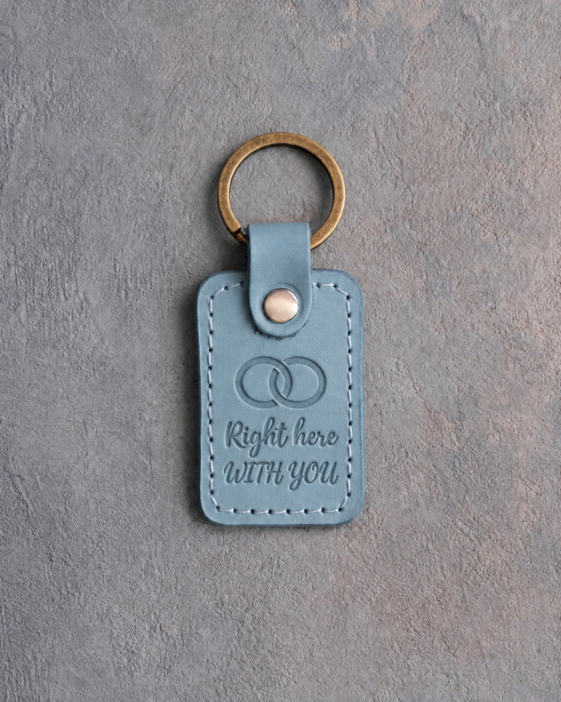 Turquoise leather keychain debossed with two interlocked wedding rings above Right here and WITH YOU, on gray concrete