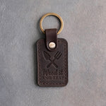 Dark espresso leather keychain debossed with a crossed trowel and hand fork above Weed it AND REAP, on gray concrete