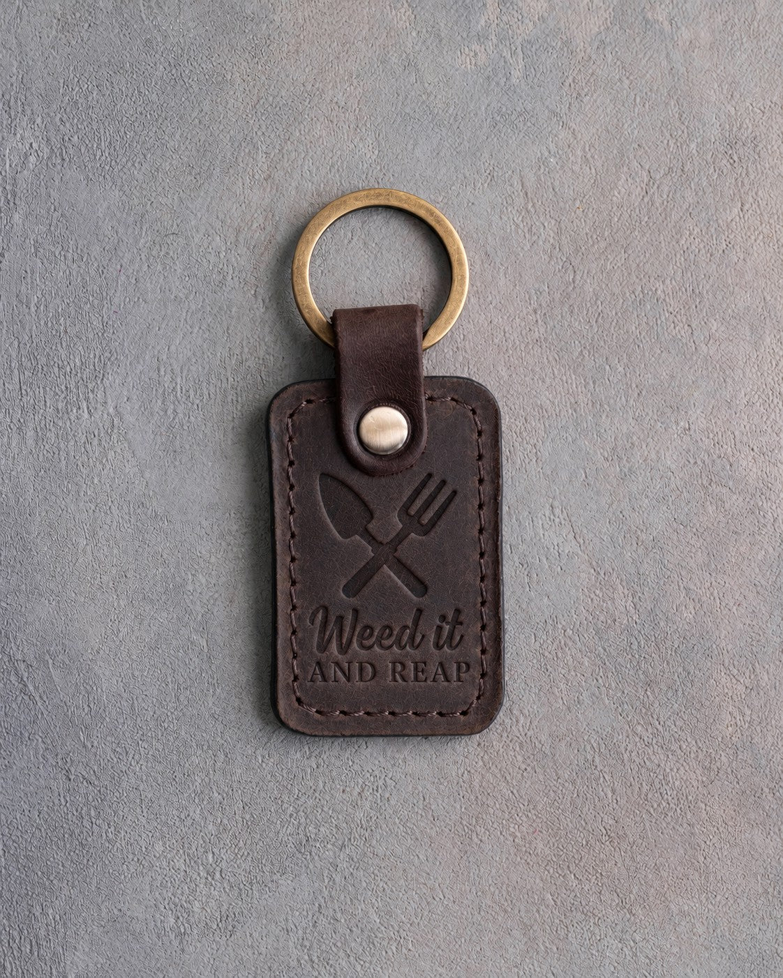 Dark espresso leather keychain debossed with a crossed trowel and hand fork above Weed it AND REAP, on gray concrete
