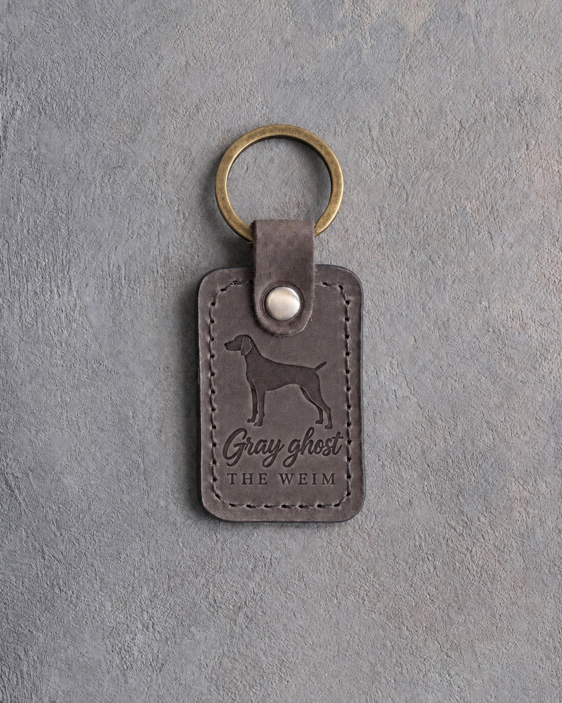 Country gray leather keychain debossed with a weimaraner above Gray ghost and THE WEIM, on gray concrete