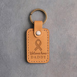Golden yellow leather keychain debossed with a looped ribbon above the words Welcome home DADDY, on gray concrete