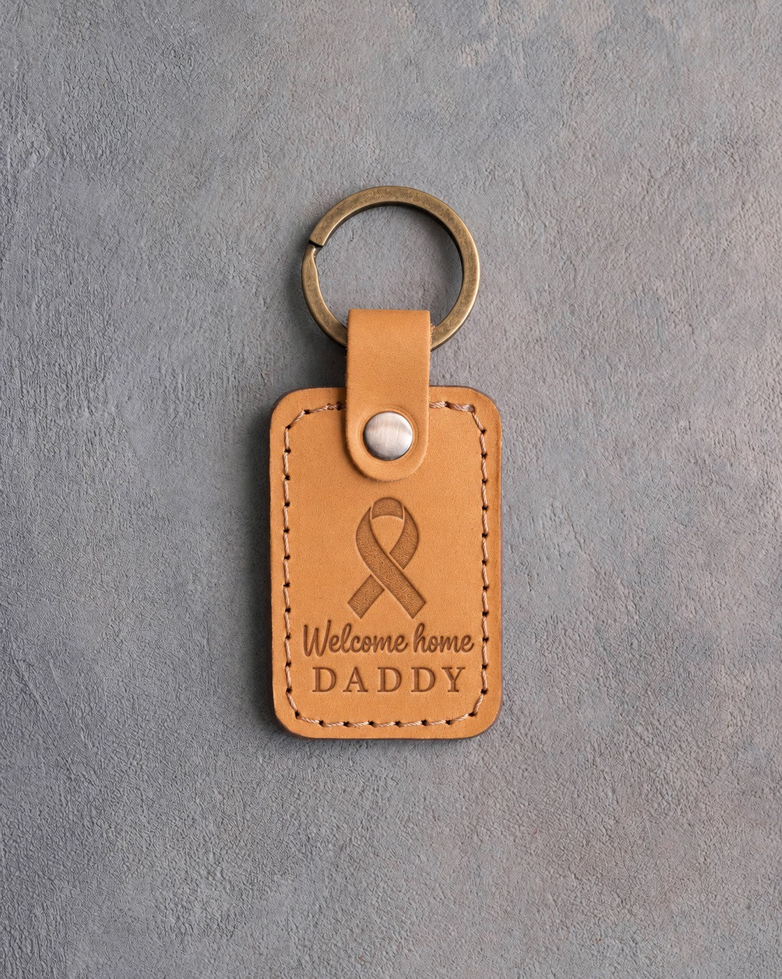 Golden yellow leather keychain debossed with a looped ribbon above the words Welcome home DADDY, on gray concrete