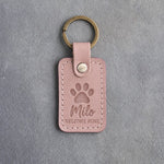 Blush pink leather keychain debossed with a single paw print above Milo and WELCOME HOME, on gray concrete
