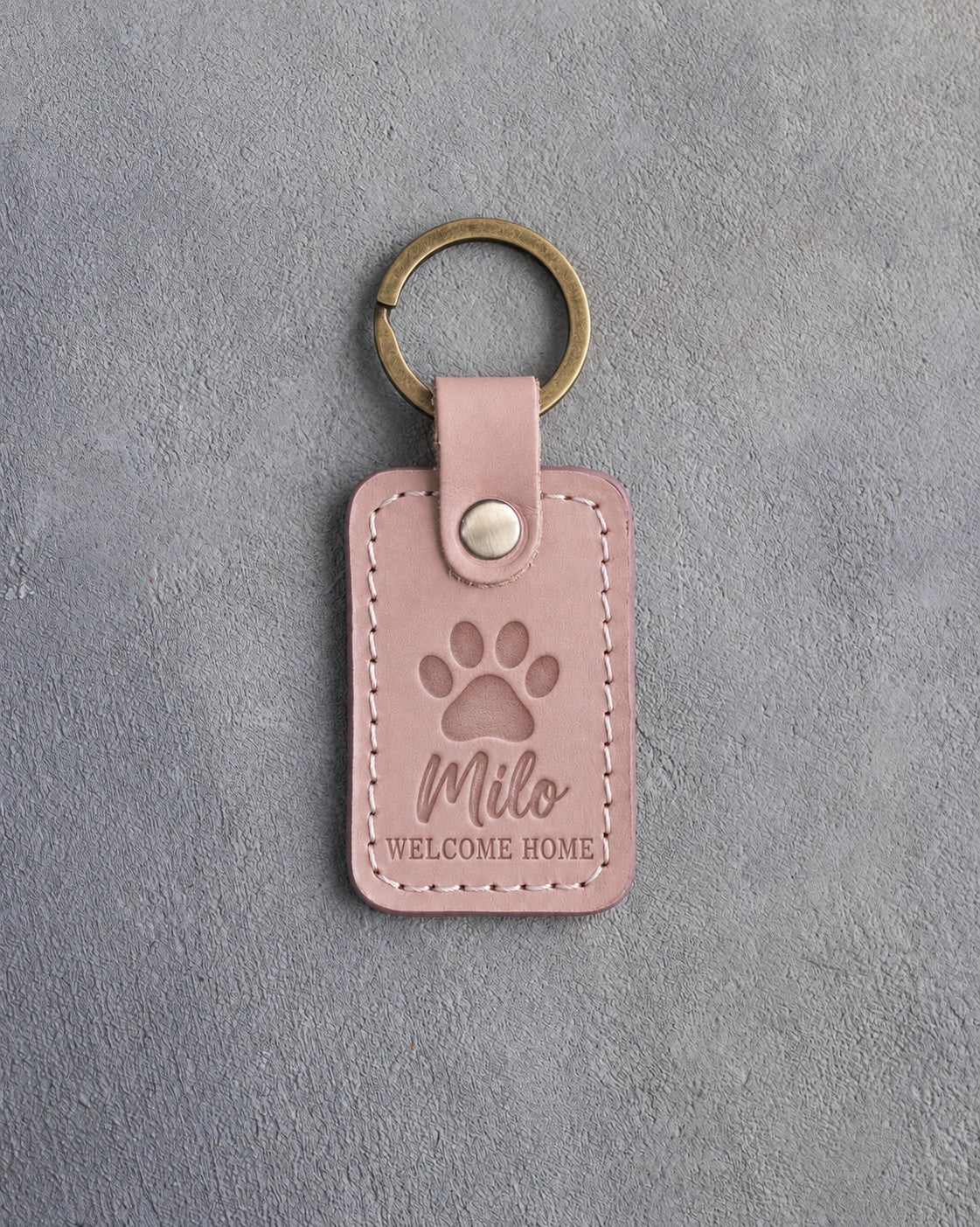 Blush pink leather keychain debossed with a single paw print above Milo and WELCOME HOME, on gray concrete
