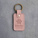 Blush pink leather keychain debossed with a single paw print above Welcome home and LITTLE ONE, on gray concrete