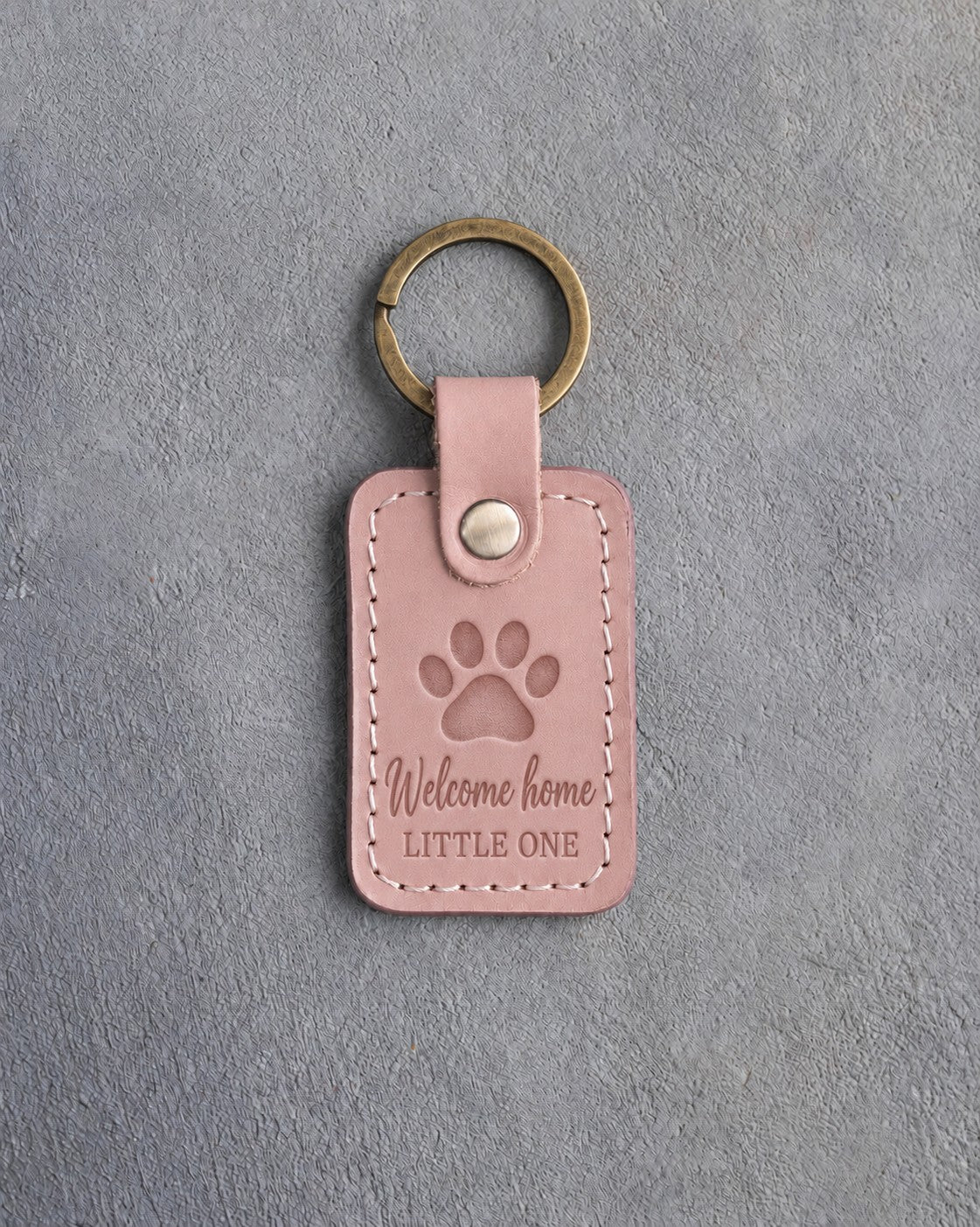 Blush pink leather keychain debossed with a single paw print above Welcome home and LITTLE ONE, on gray concrete