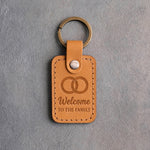 Indian Summer golden tan leather keychain debossed with two interlocked wedding rings above Welcome TO THE FAMILY, on gray concrete