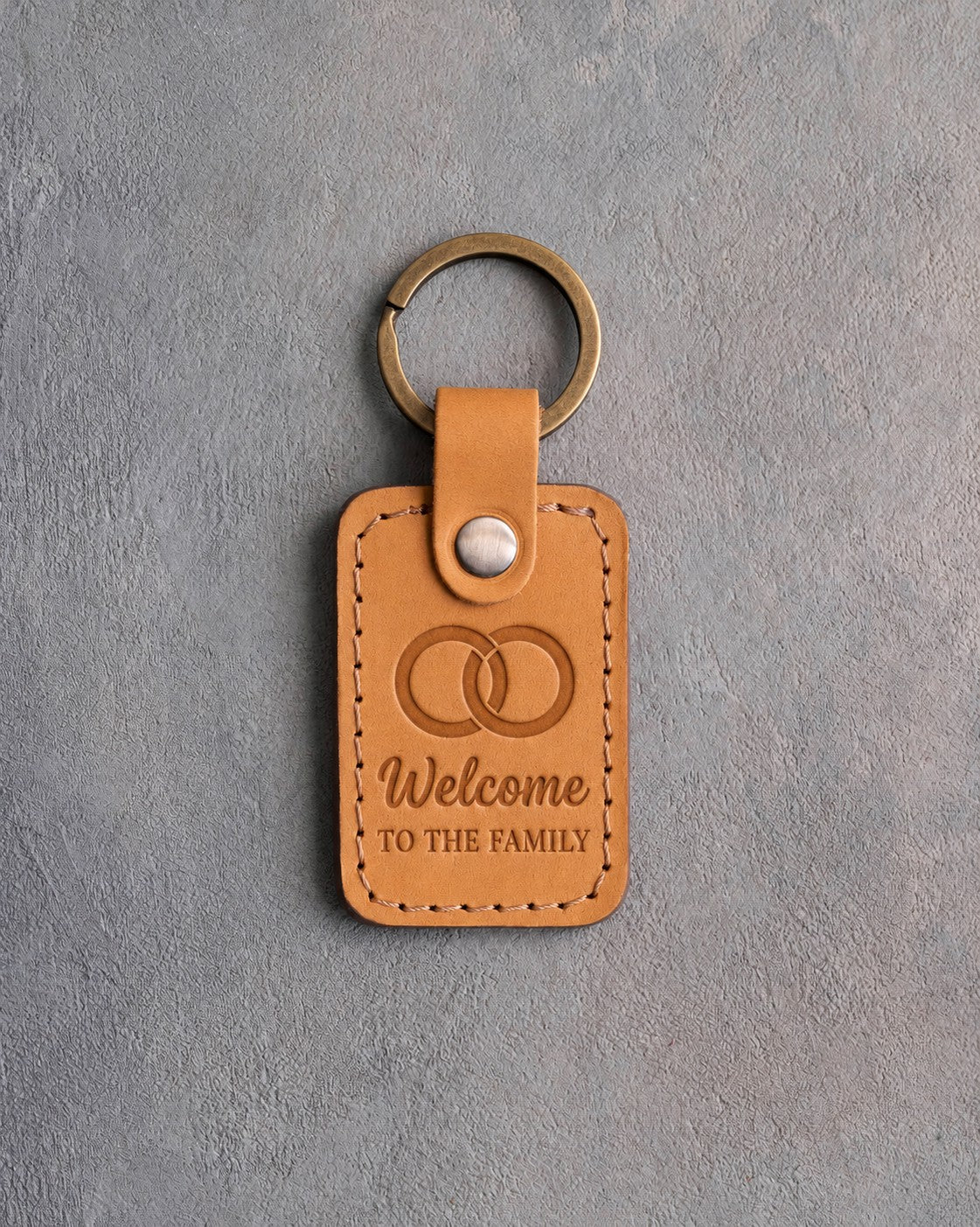 Indian Summer golden tan leather keychain debossed with two interlocked wedding rings above Welcome TO THE FAMILY, on gray concrete