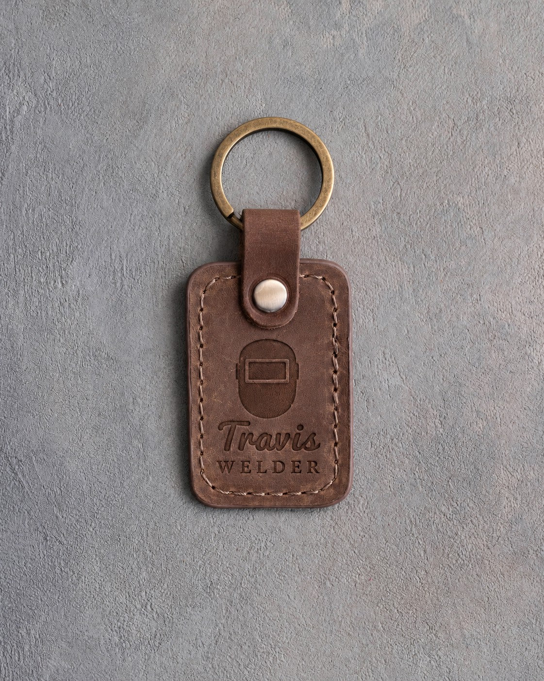 Sicilian brown leather keychain debossed with a front-view welding hood above the name Travis and WELDER, on gray plaster