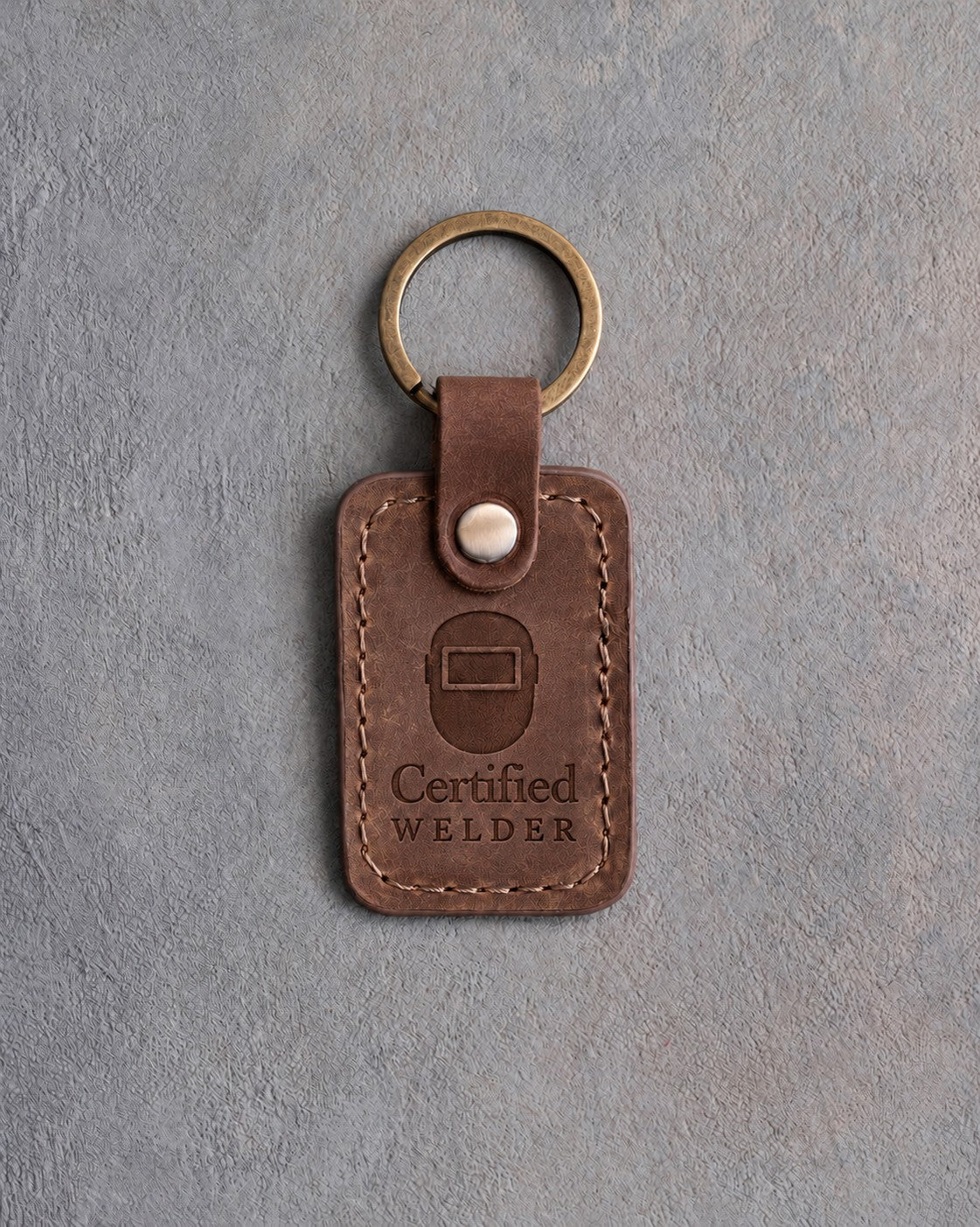 Sicilian brown leather keychain debossed with a front-view welding hood above the words Certified and WELDER, on gray concrete