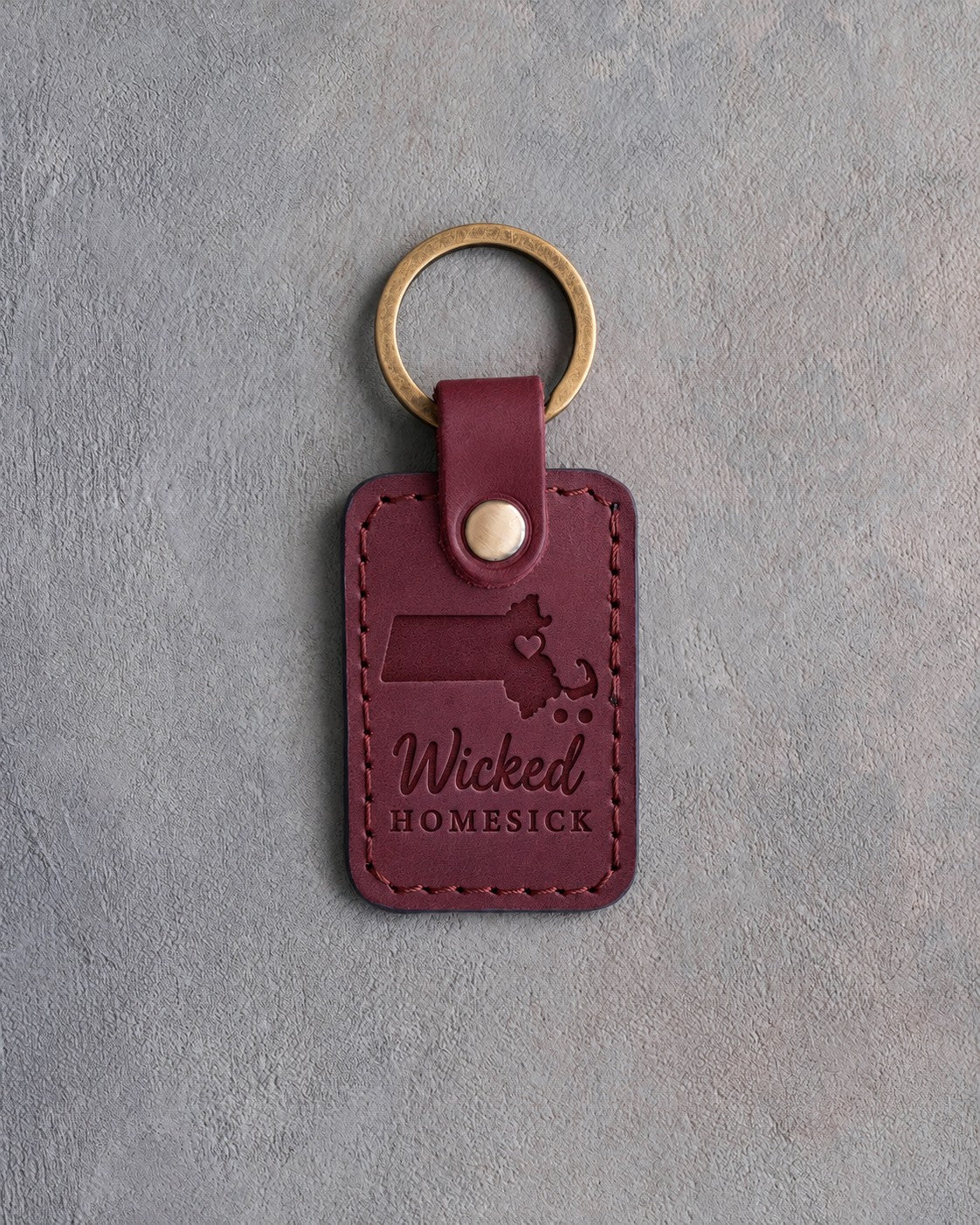Sangria red leather keychain debossed with a Massachusetts silhouette and a heart at Boston above Wicked and HOMESICK, on gray concrete