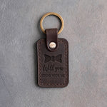Dark espresso leather keychain debossed with a bow tie above the words Will you STAND WITH ME, on gray concrete
