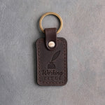 Dark espresso leather keychain debossed with a quill in an inkwell above Working and TITLE, on gray concrete