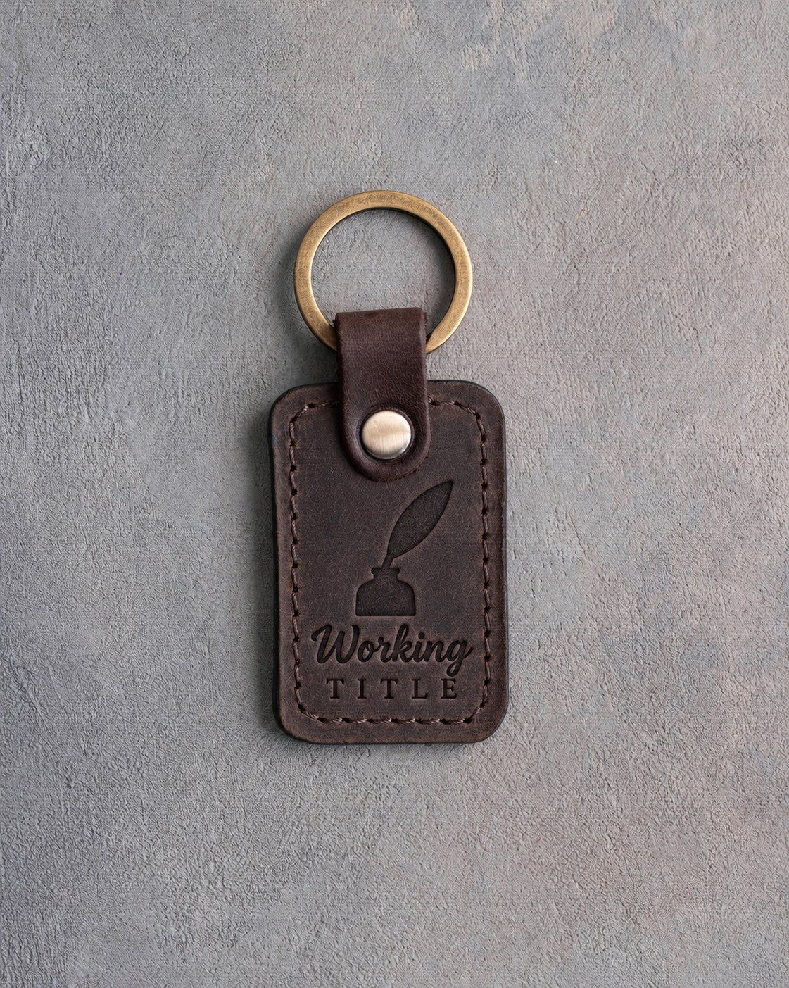Dark espresso leather keychain debossed with a quill in an inkwell above Working and TITLE, on gray concrete
