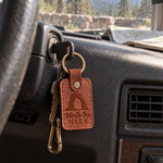 Tan Worth the Hike keychain clipped to a hiking pack resting on red slickrock at sunset