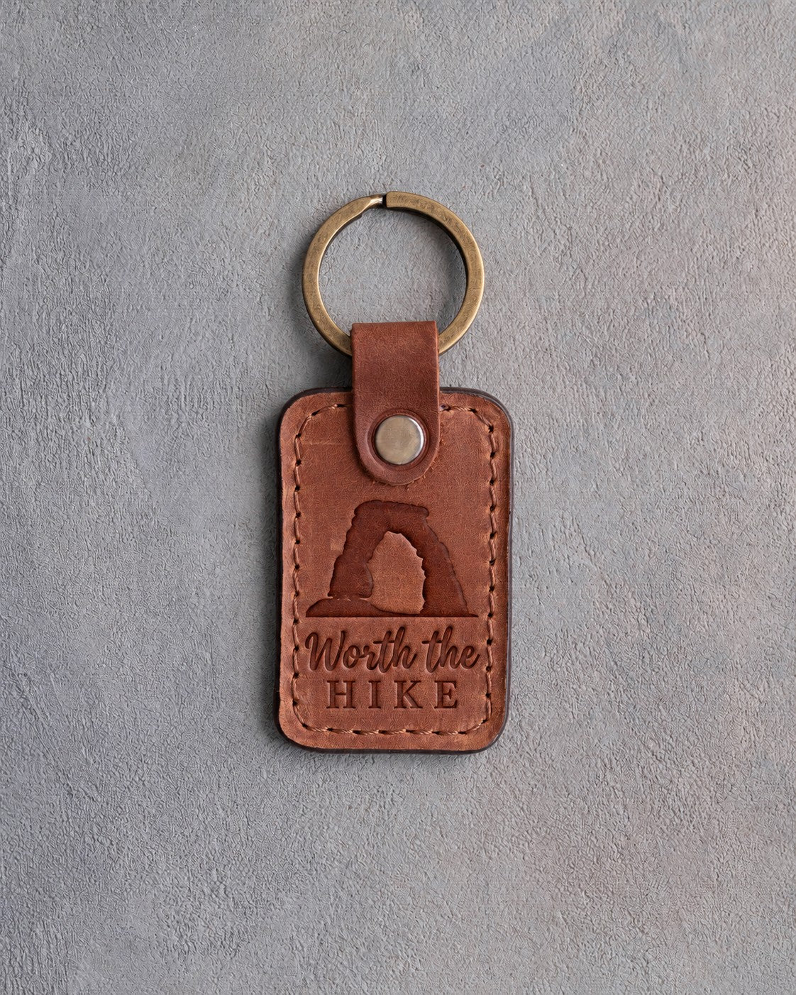 Sandy tan leather keychain debossed with a sandstone arch above Worth the and HIKE, on gray concrete