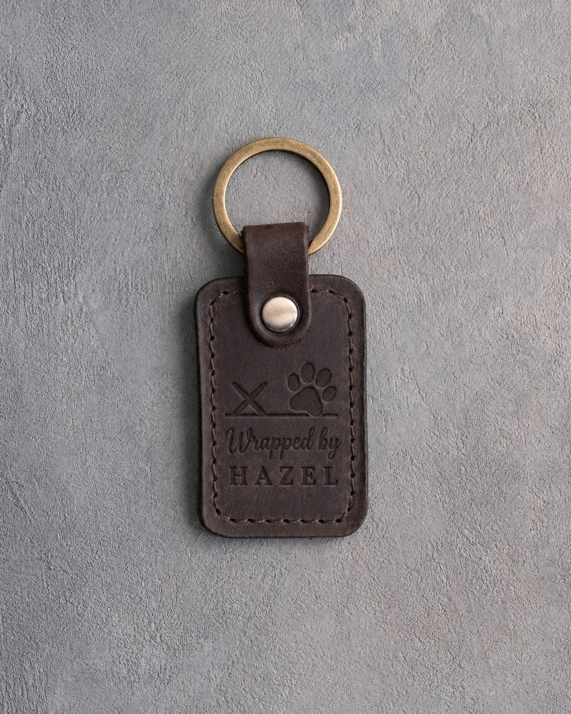 Dark espresso leather keychain debossed with a paw print on a signature line above Wrapped by and HAZEL, on gray concrete