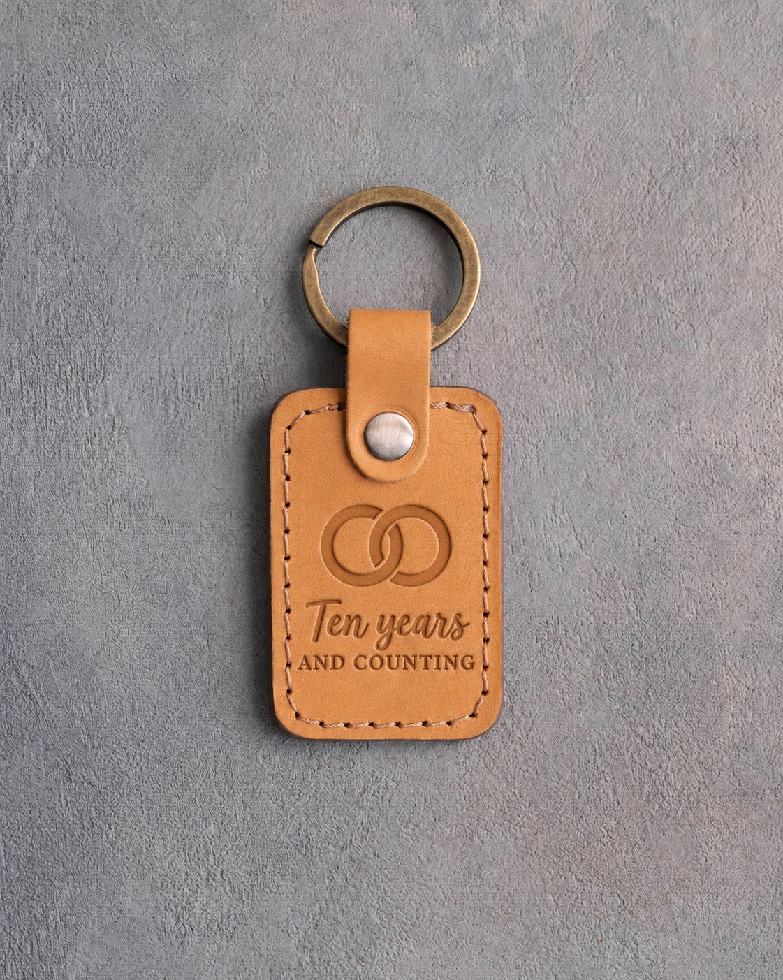 Indian Summer golden tan leather keychain debossed with two interlocked wedding rings above Ten years AND COUNTING, on gray concrete