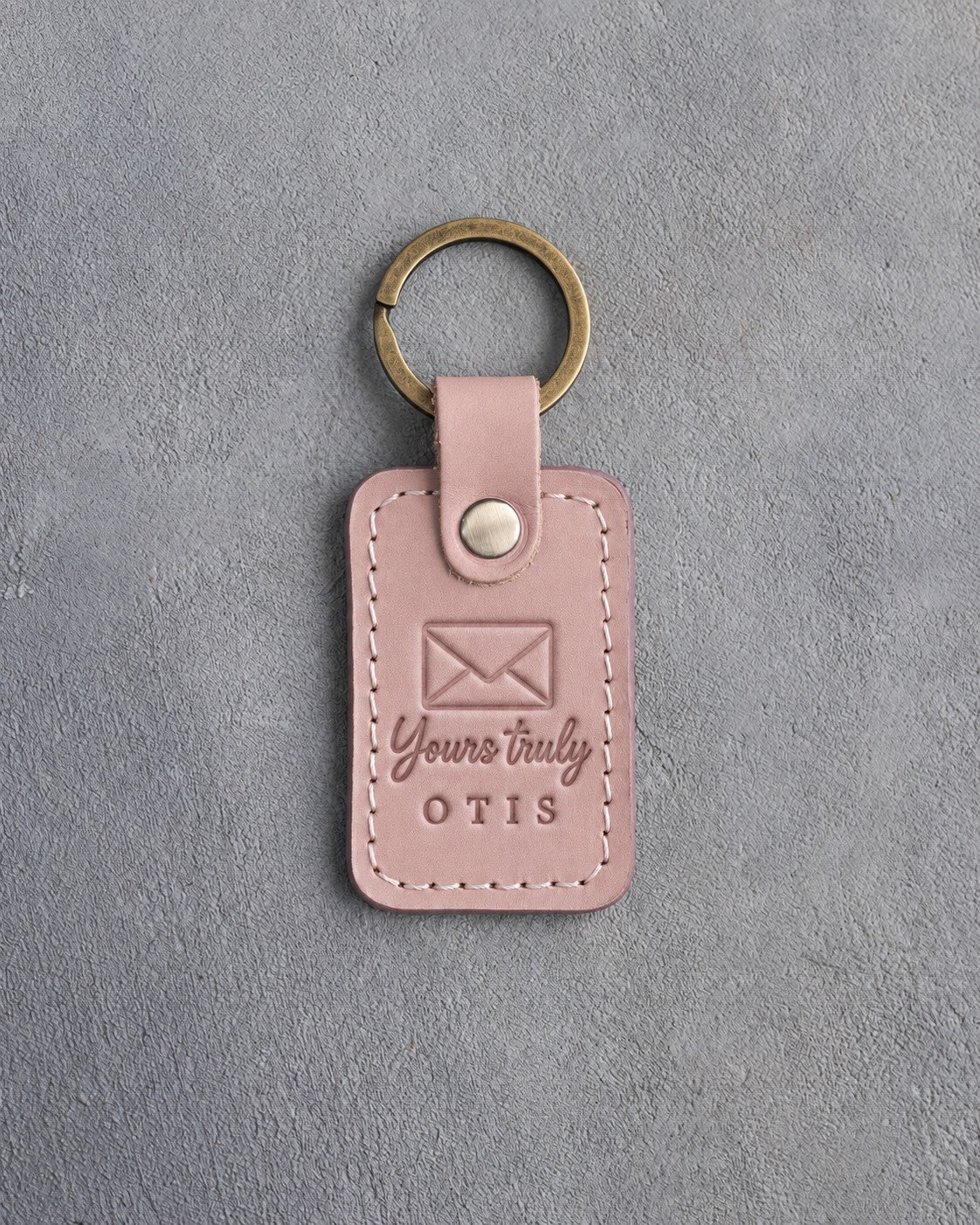 Blush pink leather keychain debossed with a sealed envelope above Yours truly and OTIS, on gray concrete