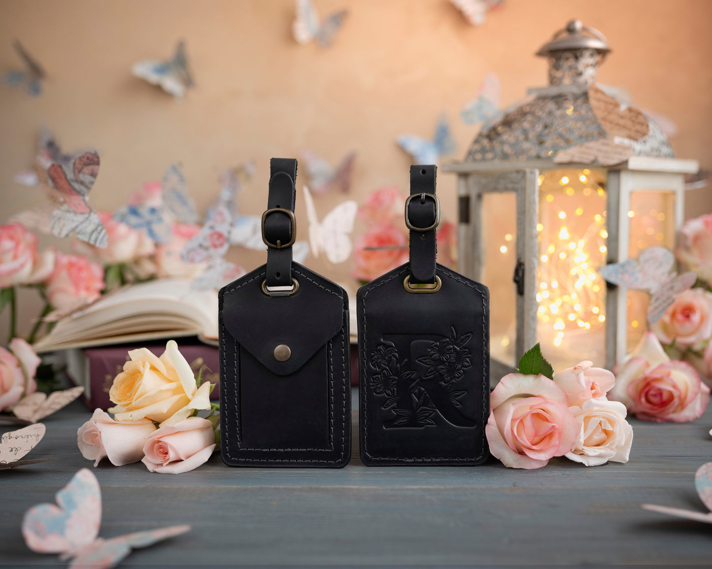 Black leather luggage tag showing the floral initial front and address-card back, staged with roses