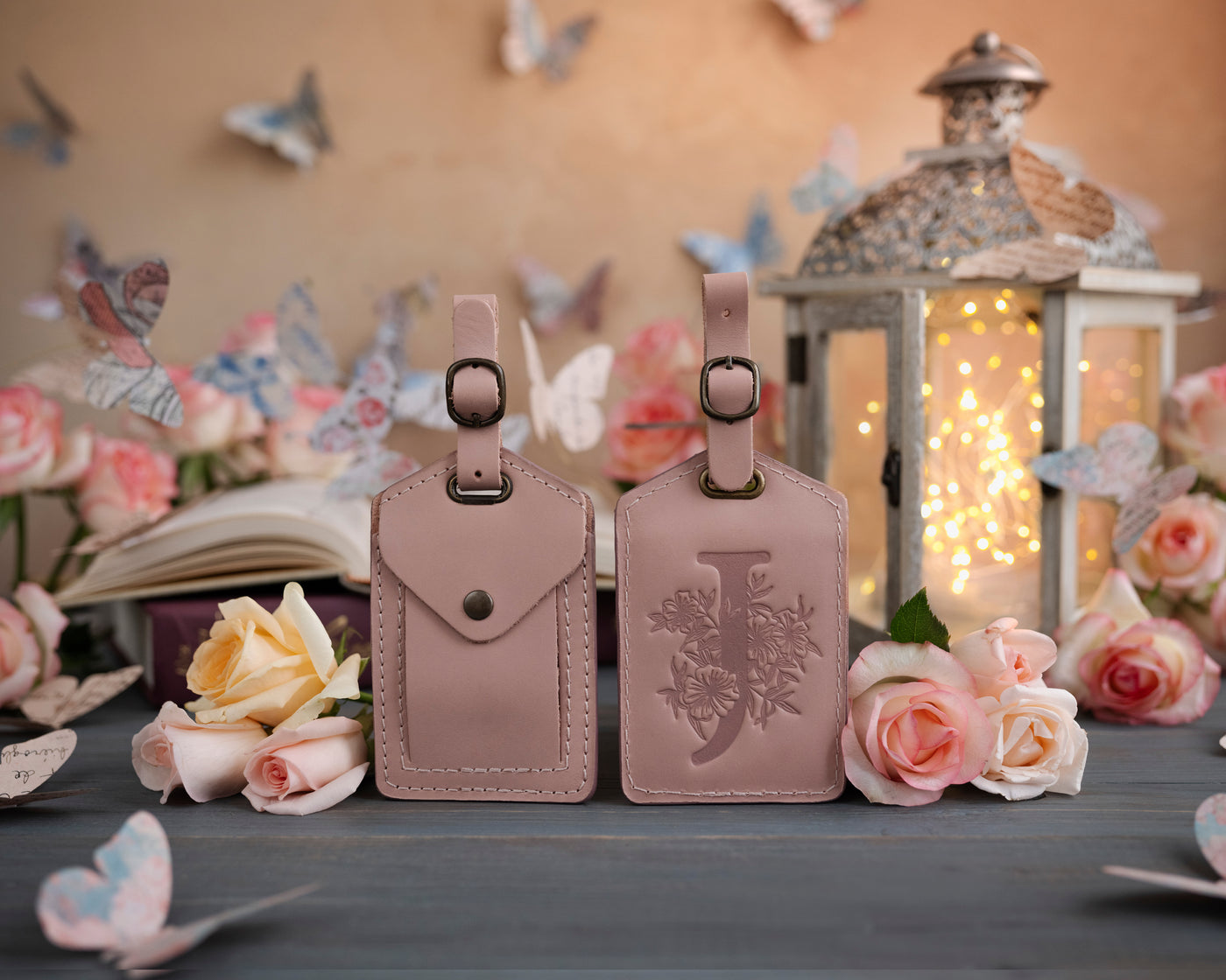Blush pink leather luggage tag showing the floral initial front and address-card back, staged with roses