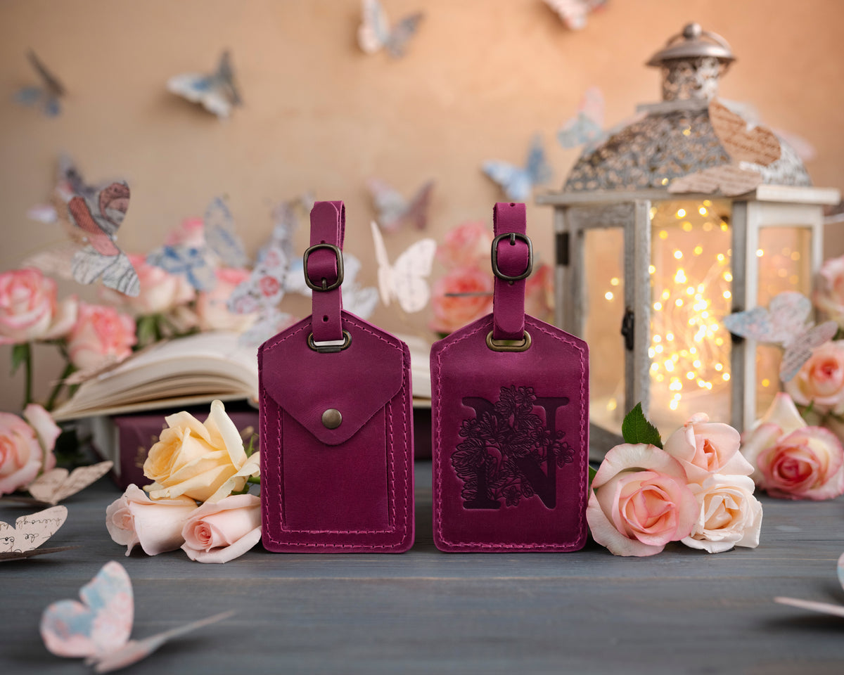 Fuchsia leather luggage tag showing the floral initial front and address-card back, staged with roses