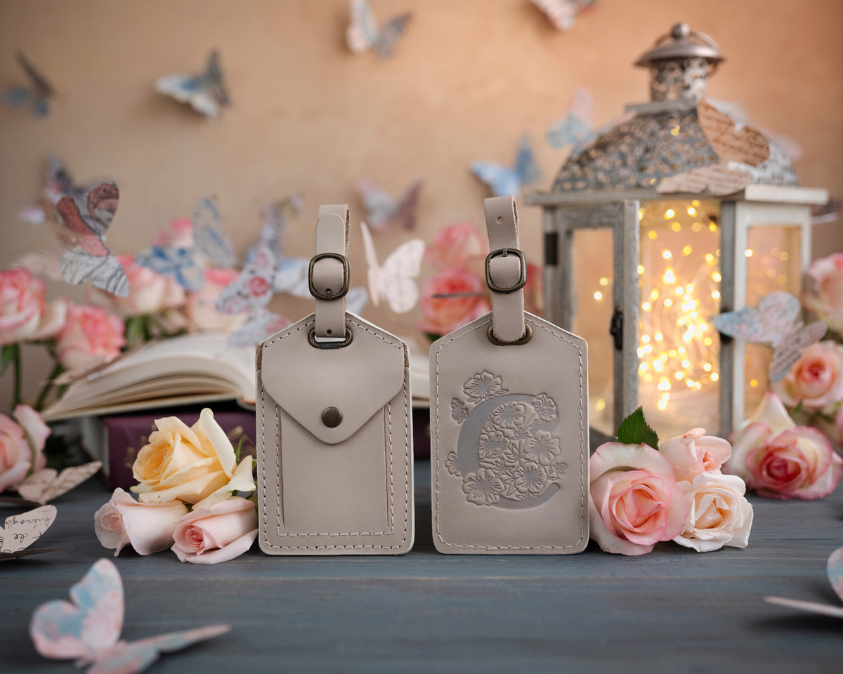 Gray sand leather luggage tag showing the floral initial front and address-card back, staged with roses