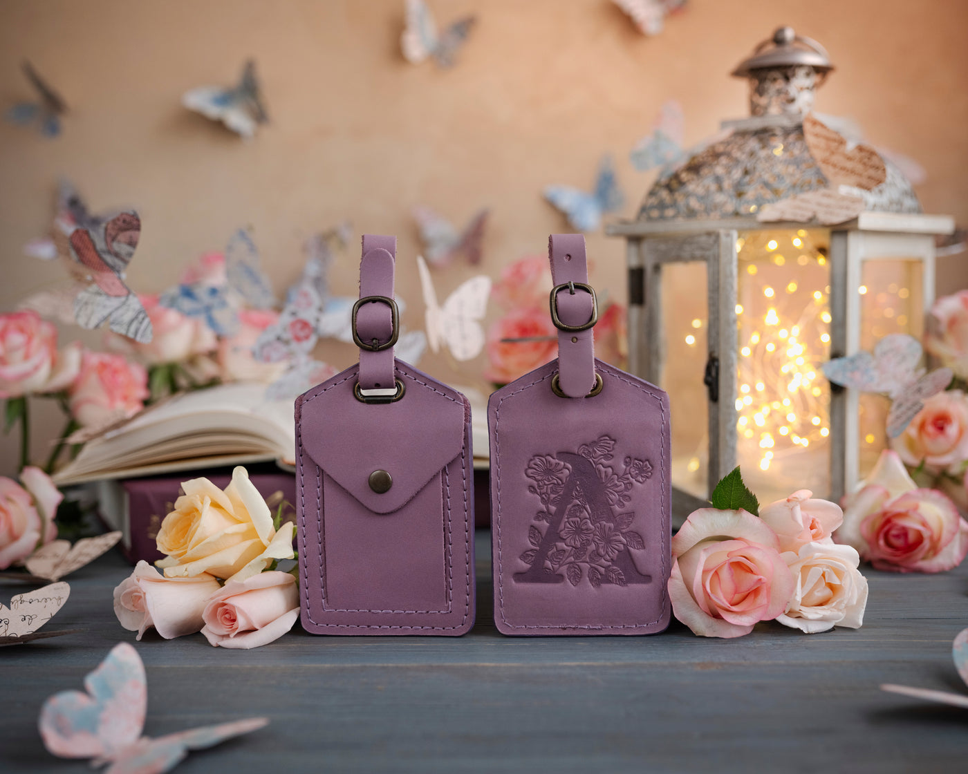 Lilac leather luggage tag showing the floral initial front and address-card back, staged with roses