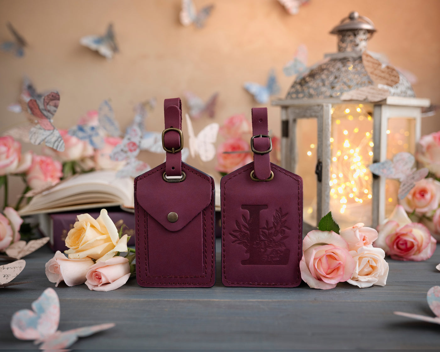 Sangria burgundy leather luggage tag showing the floral initial front and address-card back, staged with roses