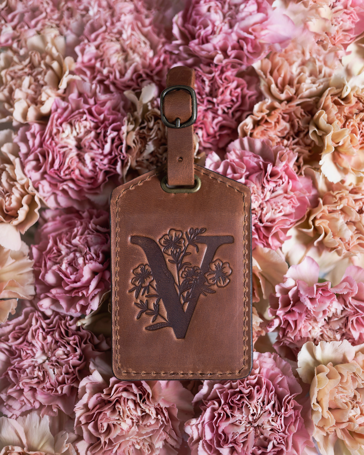 Flower initial luggage tag in caramel leather with a floral letter V deboss, resting on pink carnations