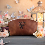 Caramel leather makeup pouch debossed with a floral letter V, set on a slate table with pale roses