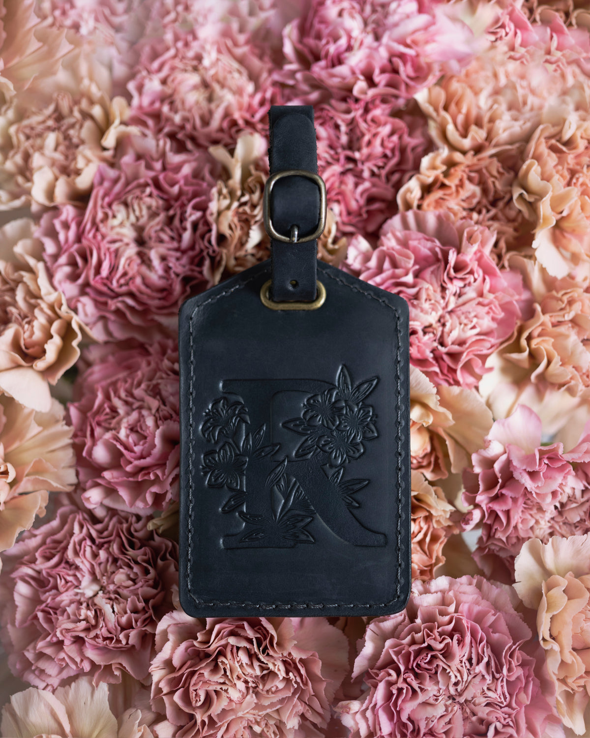Black leather luggage tag with a floral initial K and a buckle strap, on a bed of pink carnations