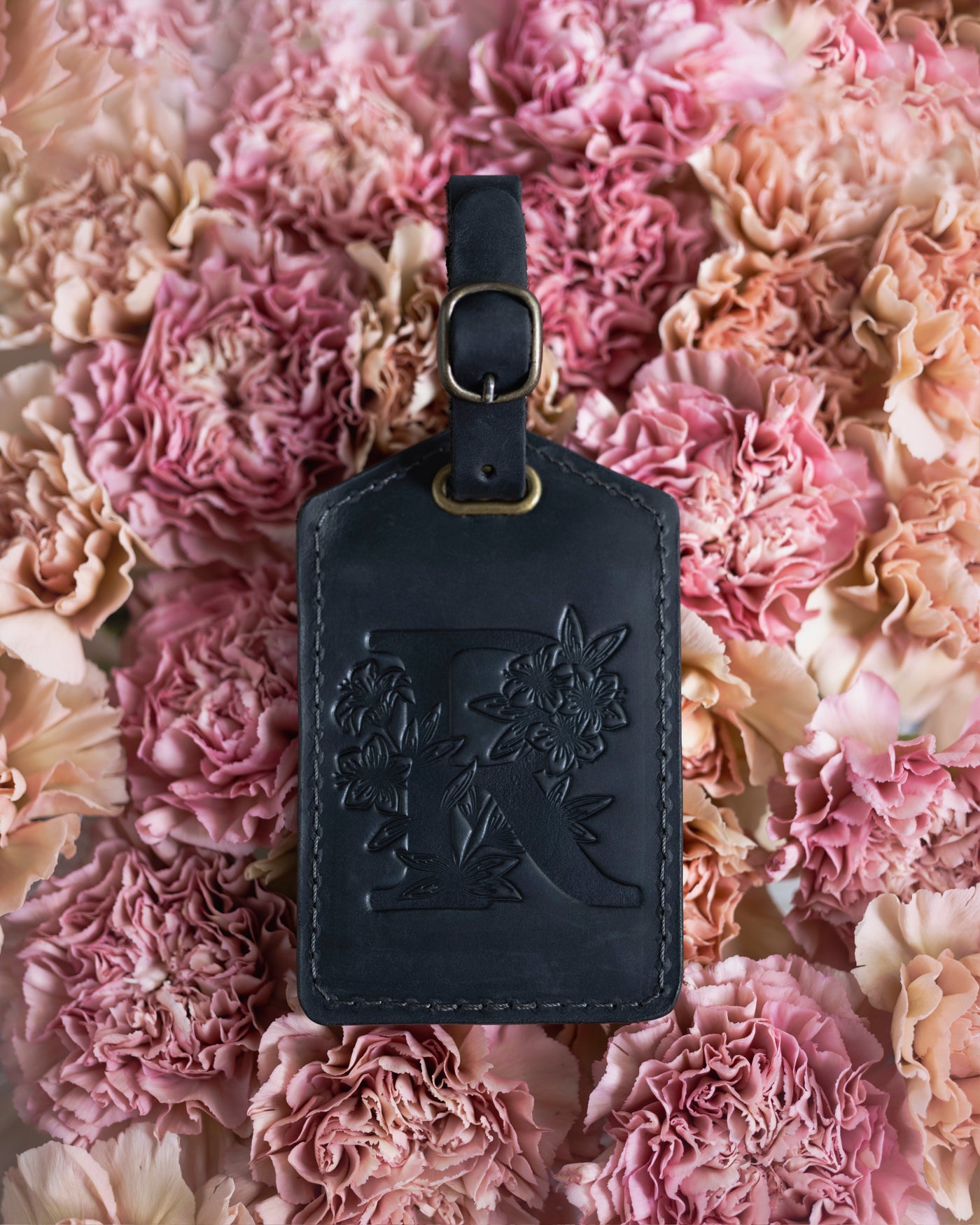 Floral initial luggage tag in black leather with a monogram deboss, on a bed of pink carnations