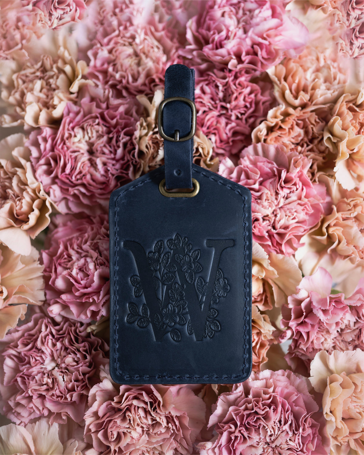 Floral initial luggage tag in blueberry navy leather with a monogram deboss, on a bed of pink carnations