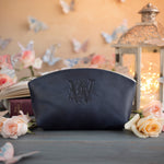 Navy blueberry leather makeup pouch stamped with a floral initial W, beside a warm lit lantern