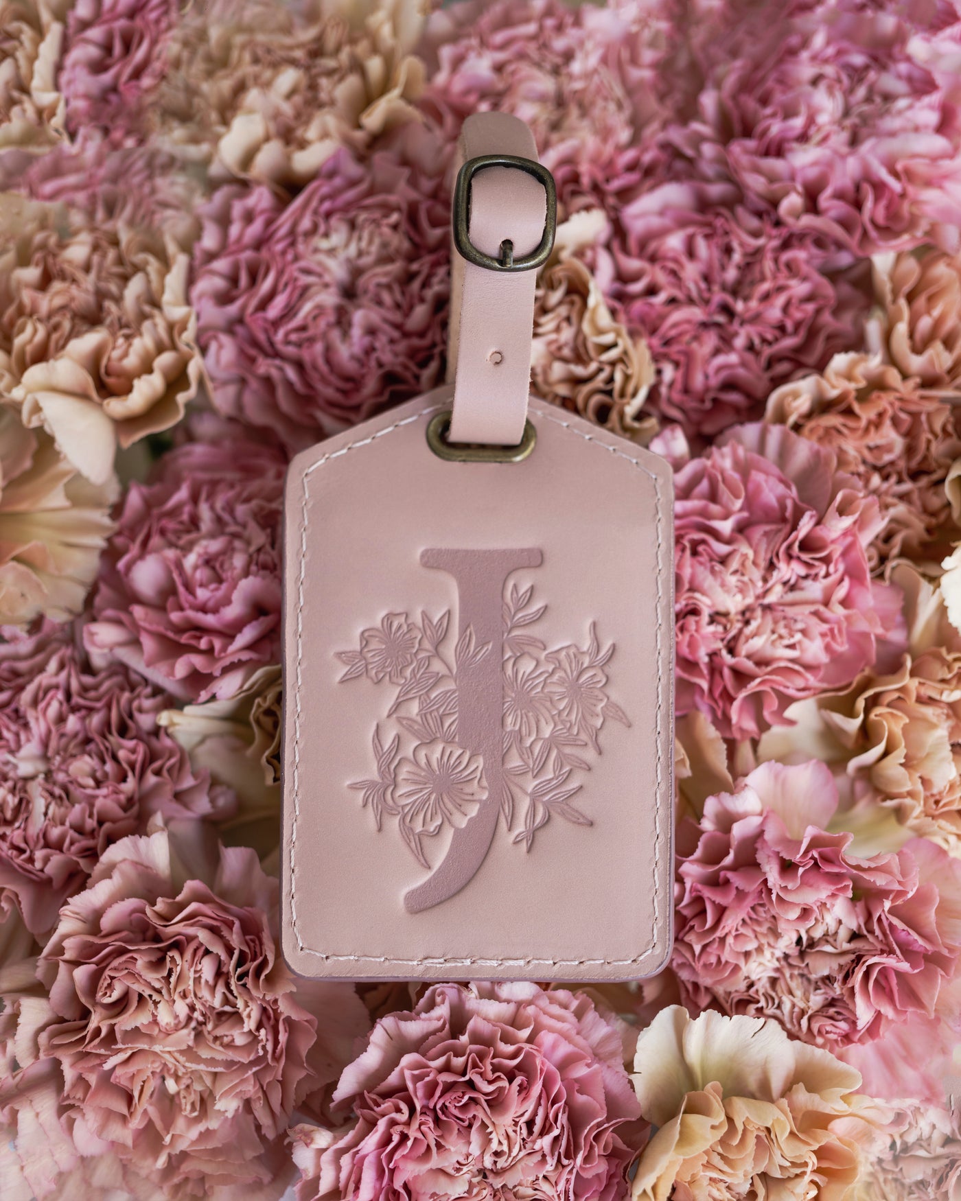 Floral initial luggage tag in blush pink leather with a monogram deboss, on a bed of pink carnations