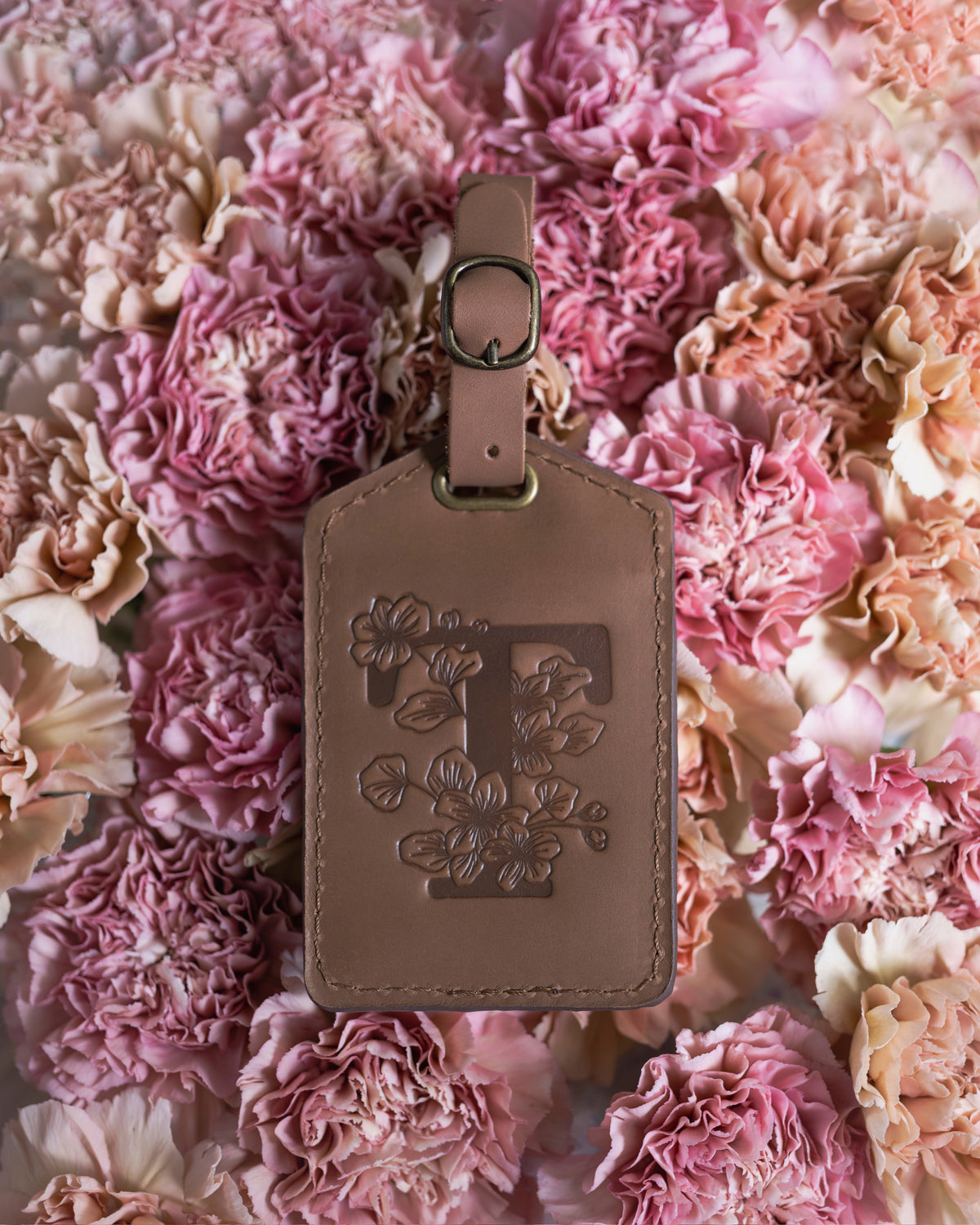 Blush tan leather luggage tag with a floral letter T, laid on soft pink carnations