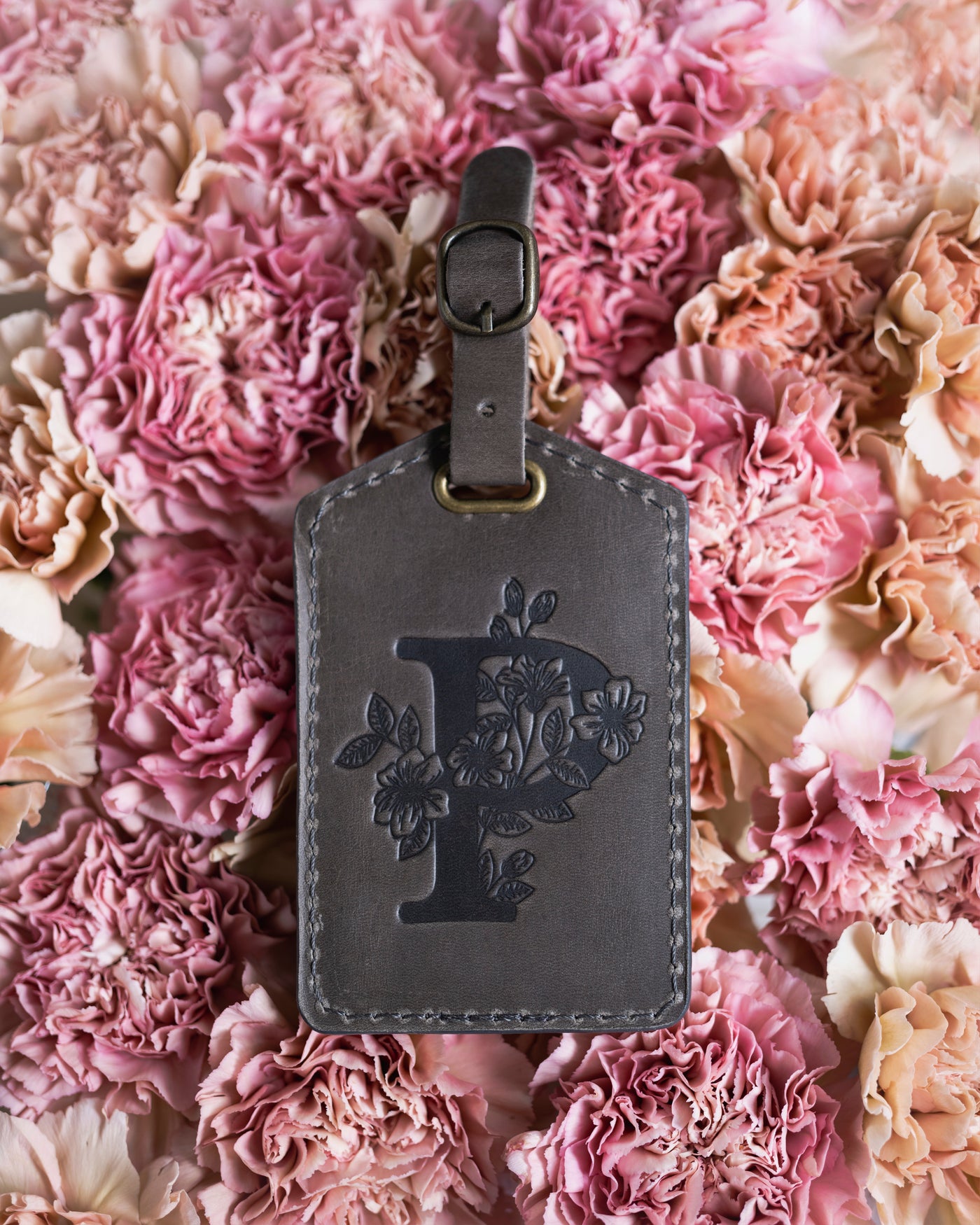 Floral initial luggage tag in country gray leather with a monogram deboss, on a bed of pink carnations