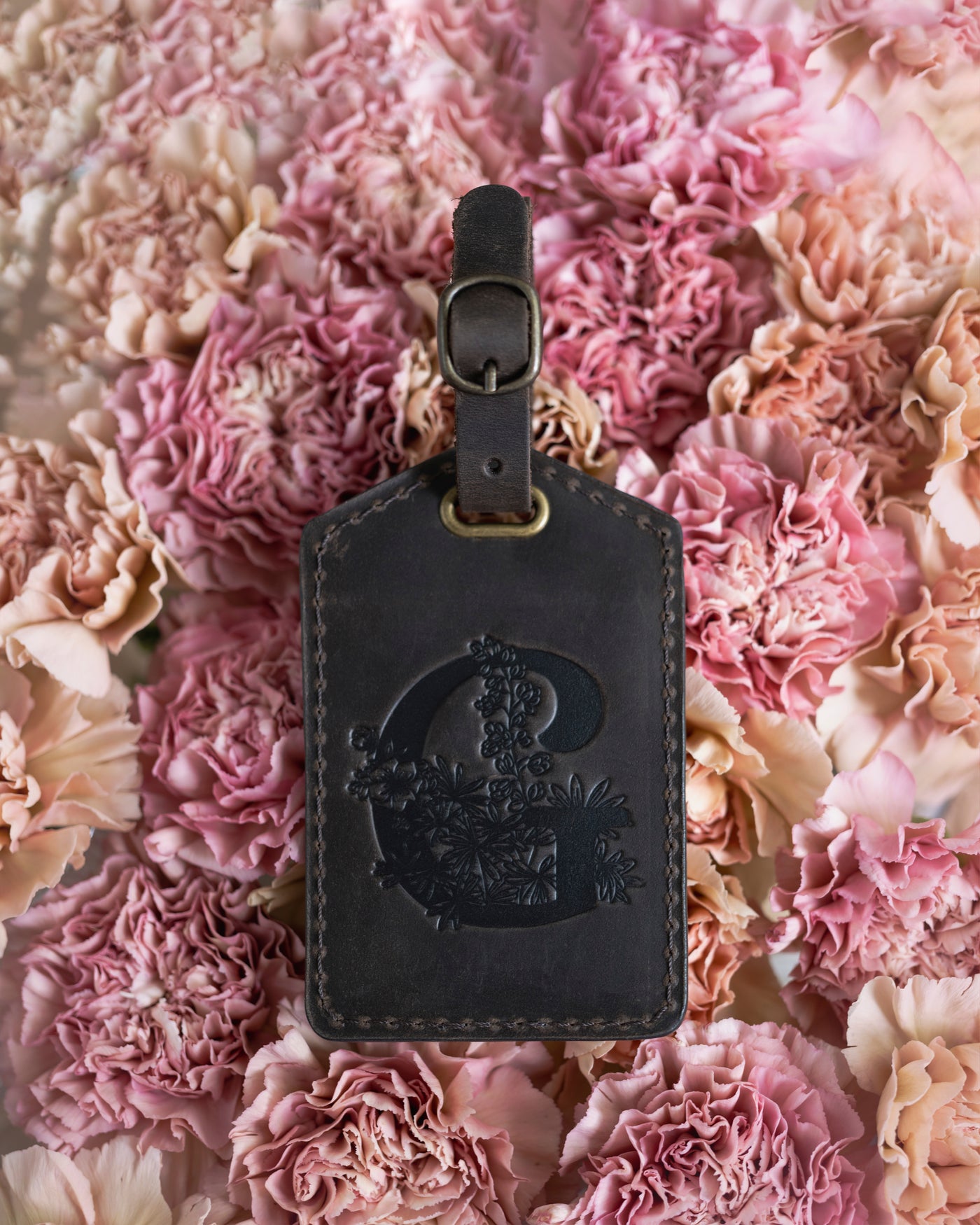 Floral initial luggage tag in dark espresso leather with a monogram deboss, on a bed of pink carnations