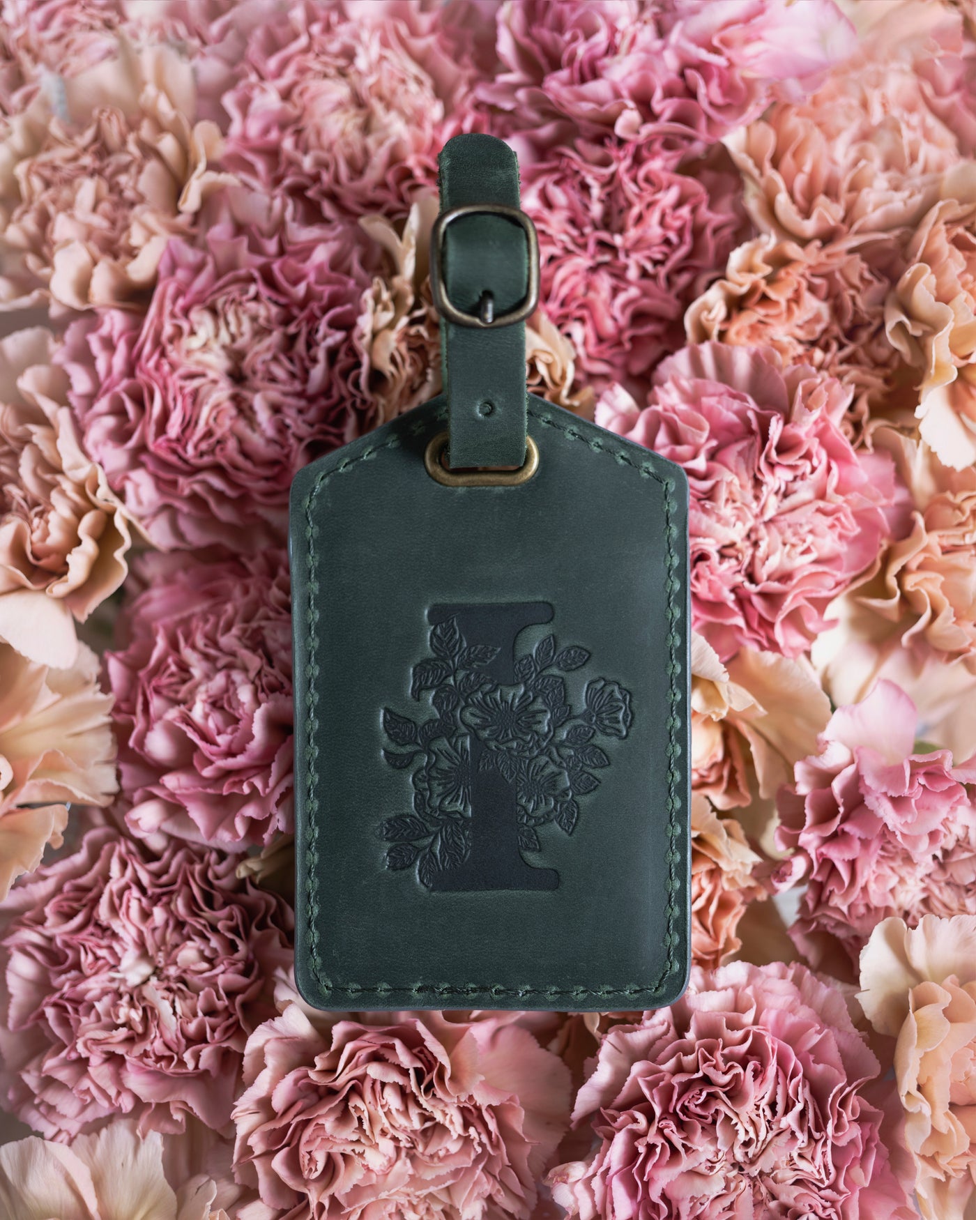 Floral initial luggage tag in forest green leather with a monogram deboss, on a bed of pink carnations