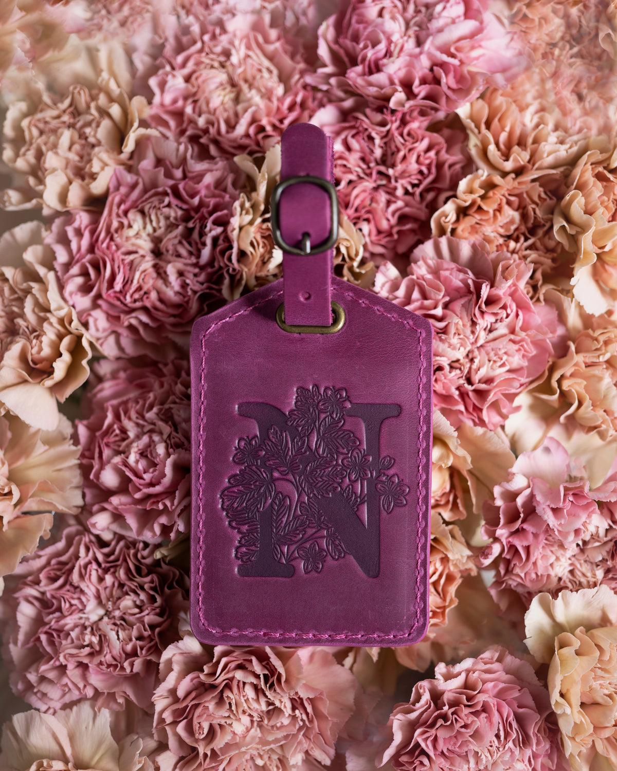 Floral initial luggage tag in fuchsia leather with a monogram deboss, on a bed of pink carnations