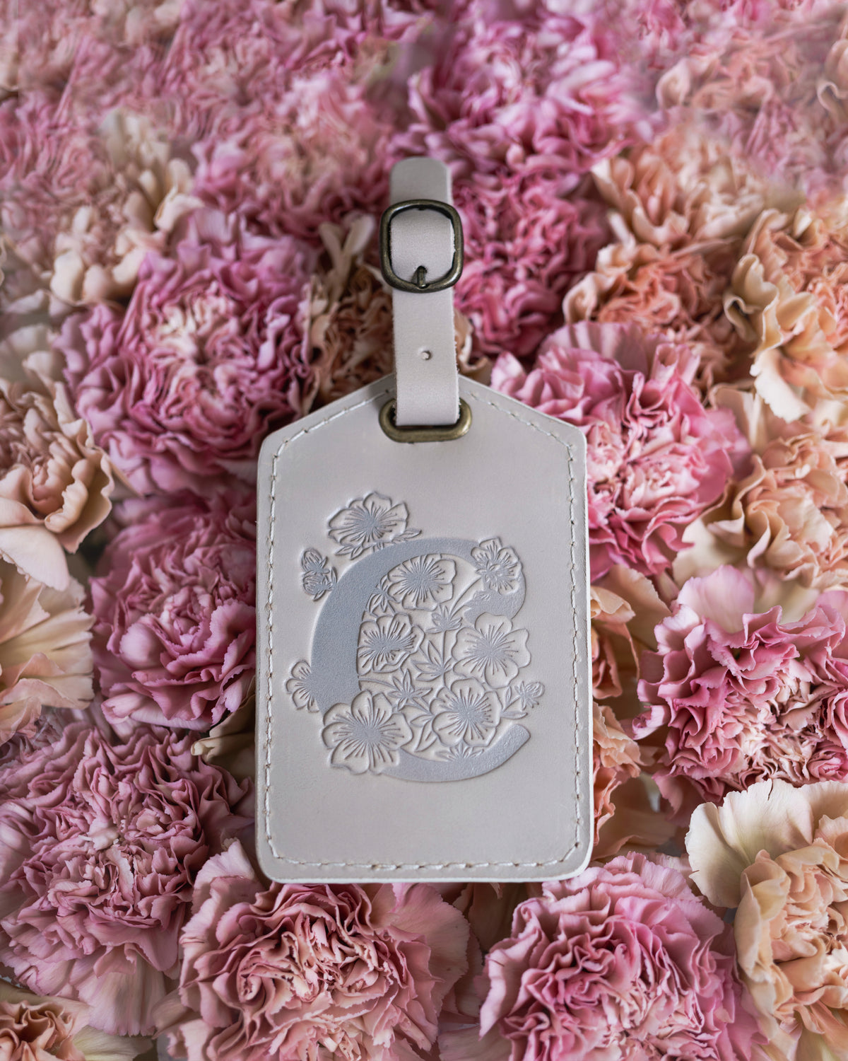 White leather luggage tag debossed with a floral letter C, resting on pink carnations