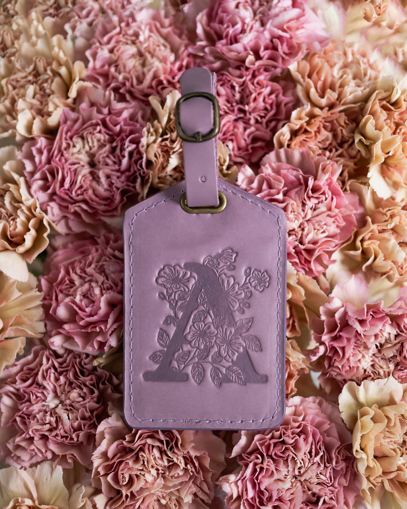 Floral initial luggage tag in lilac leather with a monogram deboss, on a bed of pink carnations
