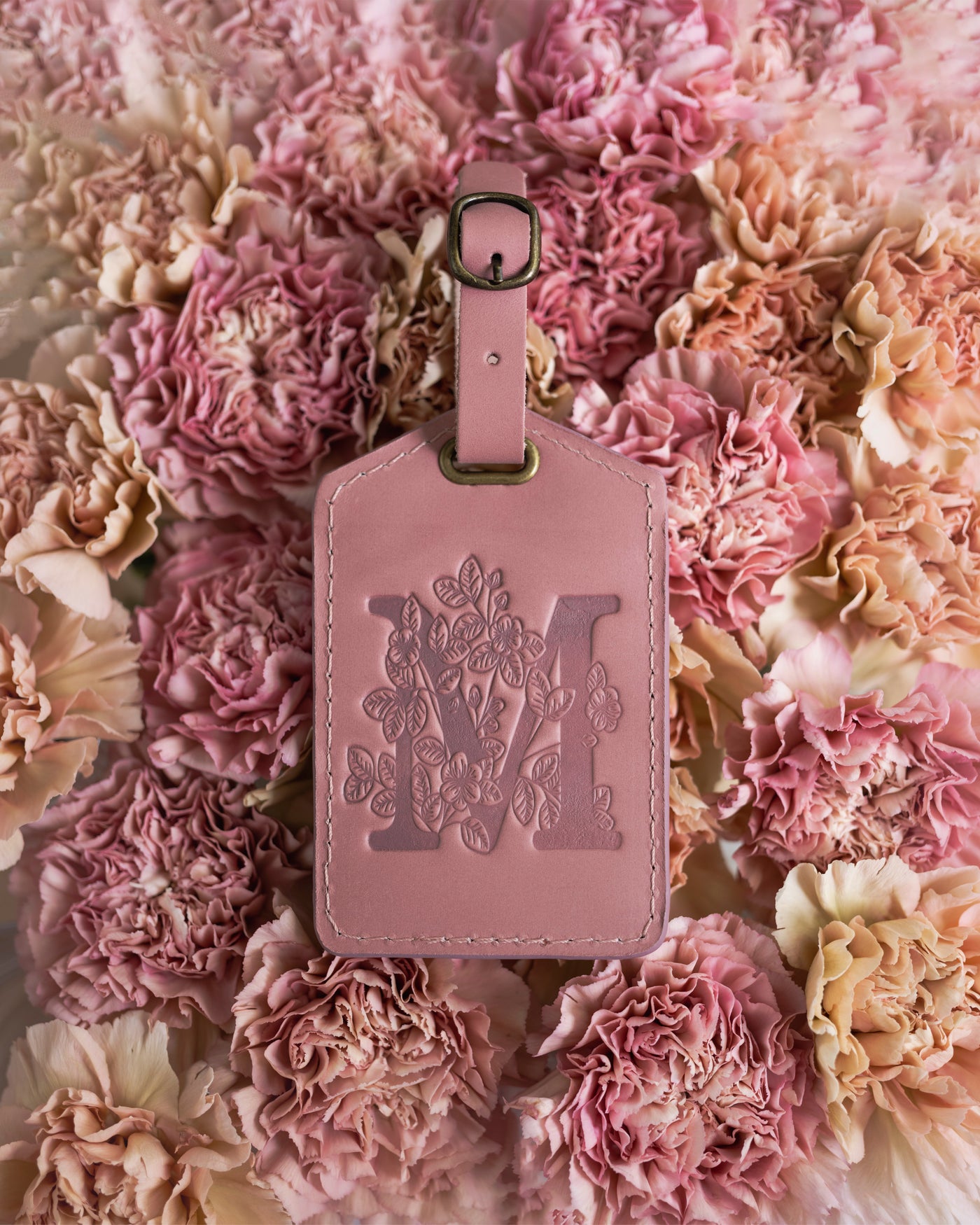 Floral initial luggage tag in parisian blossom leather with a monogram deboss, on a bed of pink carnations