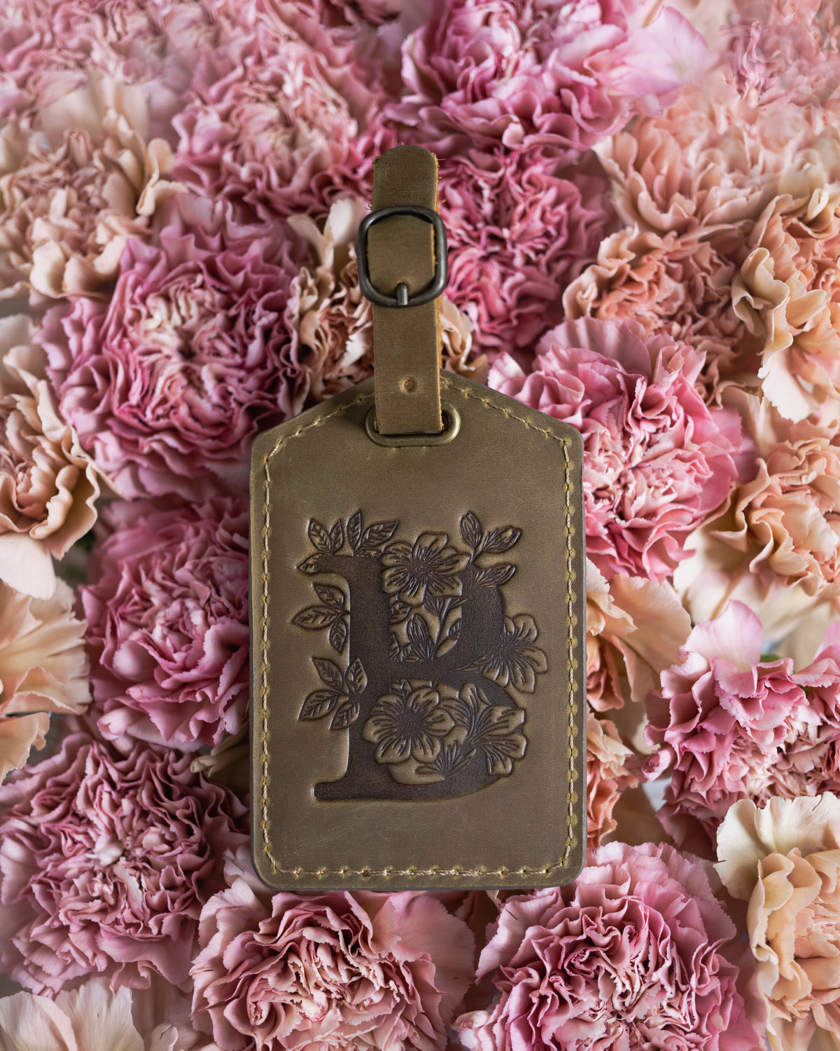 Olive arizona leather luggage tag with a floral letter L, resting on pink carnations