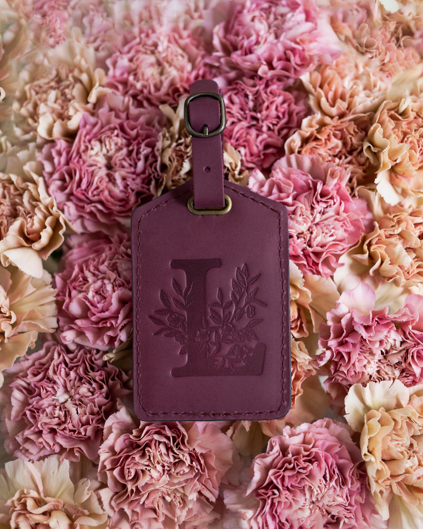 Floral initial luggage tag in sangria burgundy leather with a monogram deboss, on a bed of pink carnations