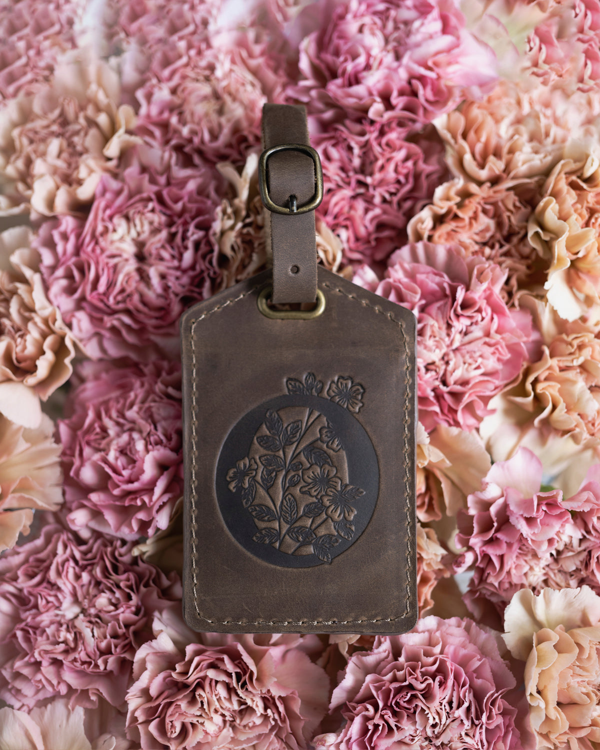 Floral initial luggage tag in sicilian brown leather with a monogram deboss, on a bed of pink carnations