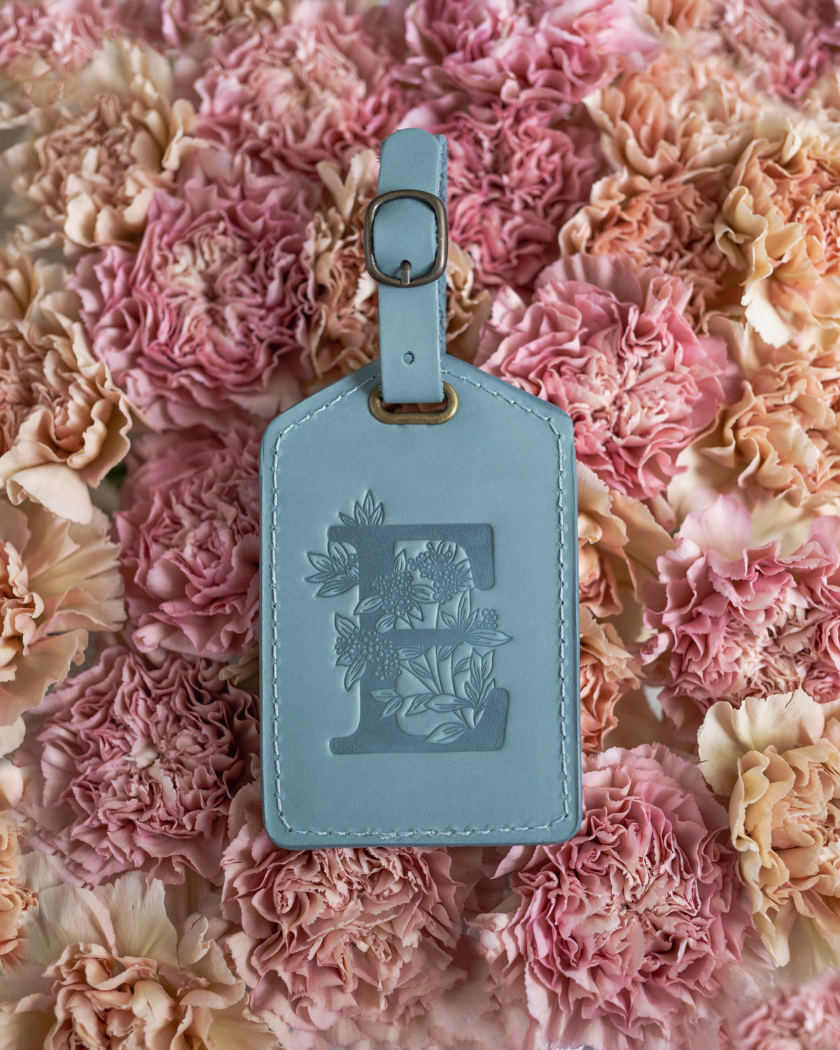 Soft blue leather luggage tag with a floral letter E, resting on pink carnations